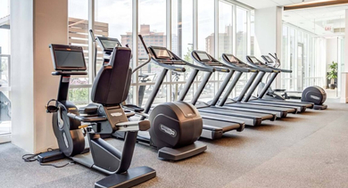 Modern fitness center with a row of treadmills, elliptical machines, and bikes, featuring large windows at North+Vine in Chicago