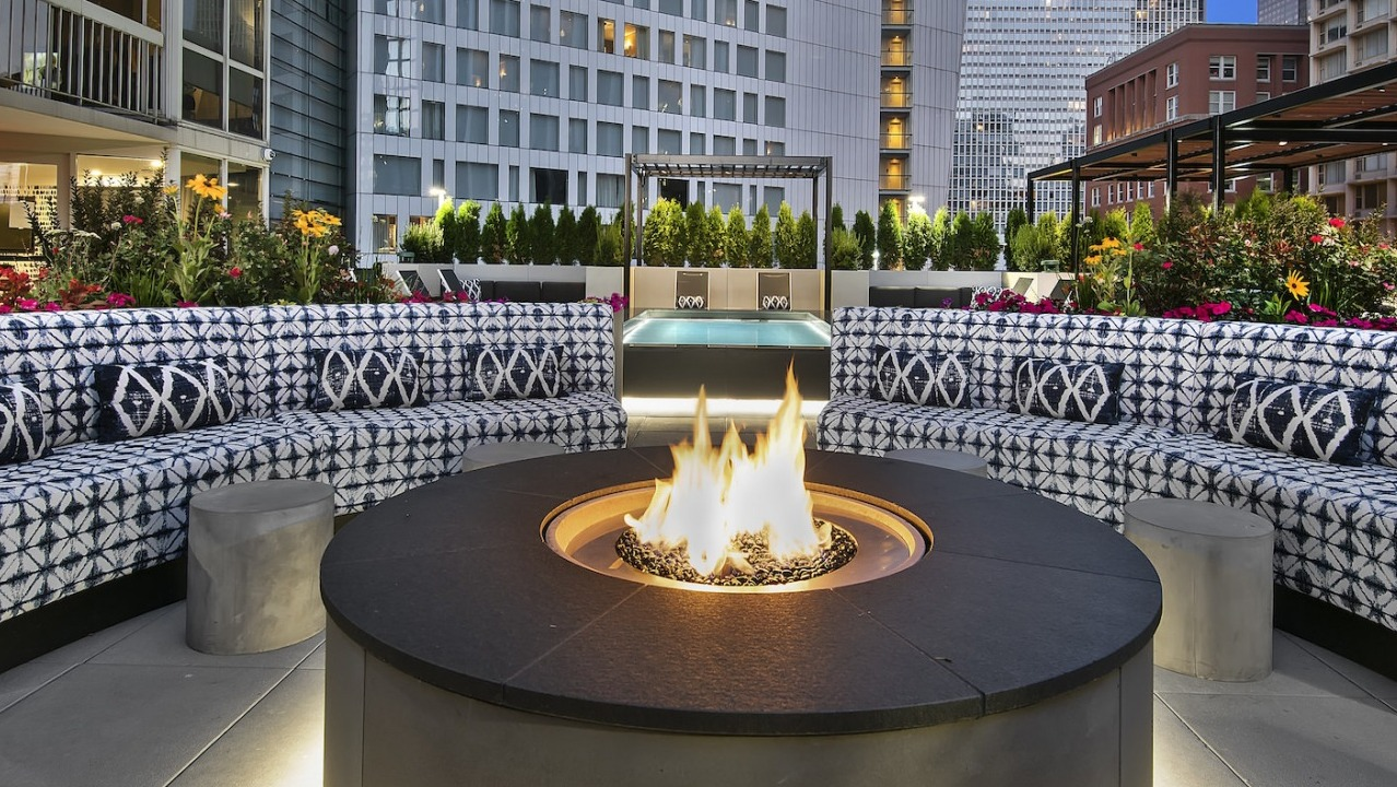 Cozy rooftop lounge area at dusk with a warm fire pit, comfortable patterned sofas, and city views at One East Delaware Apartments