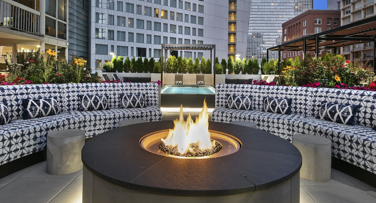 Cozy rooftop lounge area at dusk with a warm fire pit, comfortable patterned sofas, and city views at One East Delaware Apartments