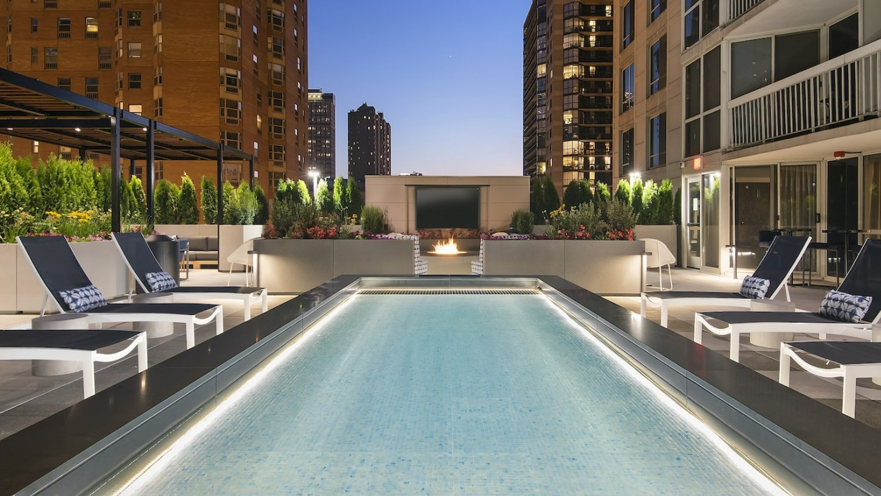 Evening view of the rooftop pool and lounge area, featuring illuminated pool, fire pit, and city lights at One East Delaware Apartments