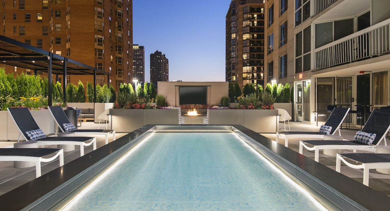 Evening view of the rooftop pool and lounge area, featuring illuminated pool, fire pit, and city lights at One East Delaware Apartments