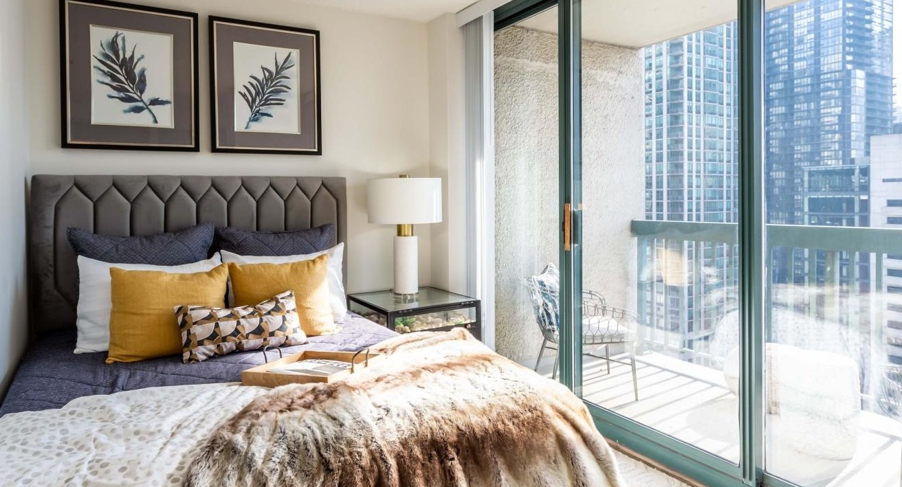Sunlit bedroom at One Superior Place Apartments featuring modern décor, warm bedding, and balcony access with high-rise city views