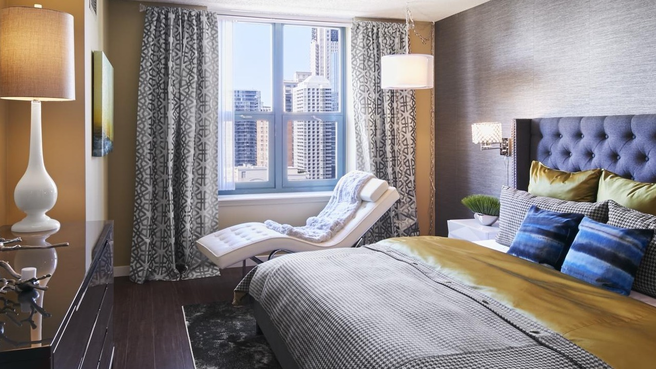 Modern bedroom at One Superior Place Apartments with tufted headboard, accent chair, and large window showcasing Chicago city views