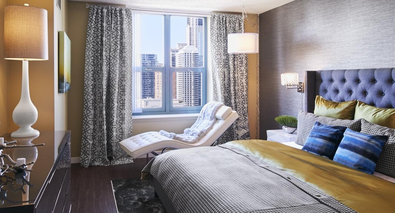 Modern bedroom at One Superior Place Apartments with tufted headboard, accent chair, and large window showcasing Chicago city views