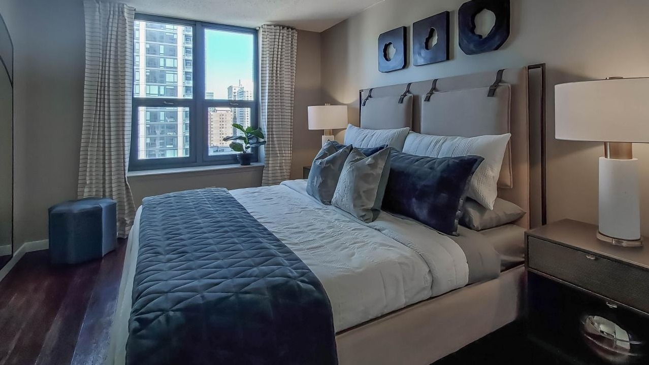 Elegant bedroom at One Superior Place Apartments with dark wood floors, upholstered bed, and decorative accents next to a large window