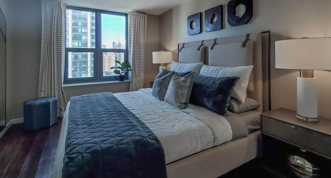 Elegant bedroom at One Superior Place Apartments with dark wood floors, upholstered bed, and decorative accents next to a large window