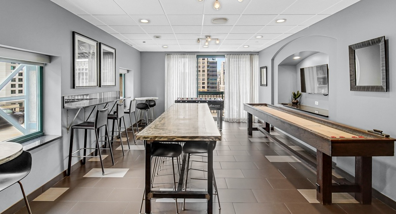 Resident game room at One Superior Place Apartments with shuffleboard table, high-top seating, and natural light from large windows