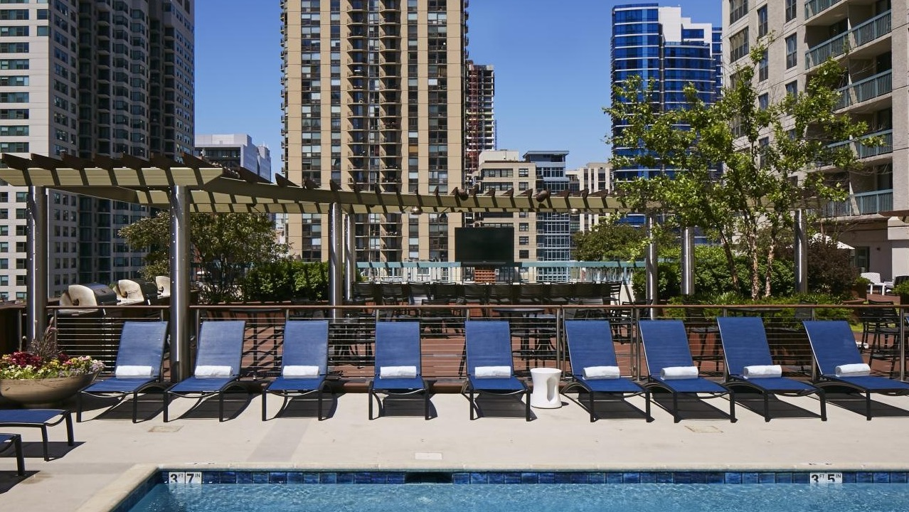 Rooftop pool and sundeck at One Superior Place Apartments featuring blue lounge chairs, pergola shade, and city high-rise views