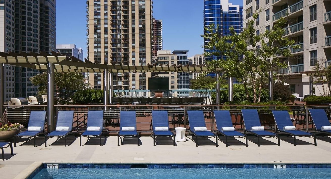Rooftop pool and sundeck at One Superior Place Apartments featuring blue lounge chairs, pergola shade, and city high-rise views