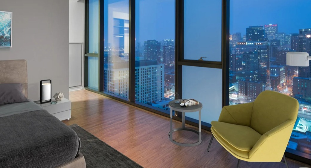 Bedroom with floor-to-ceiling windows and sweeping night skyline views at ONE333 apartments in Chicago, featuring cozy seating and modern lighting for a serene retreat