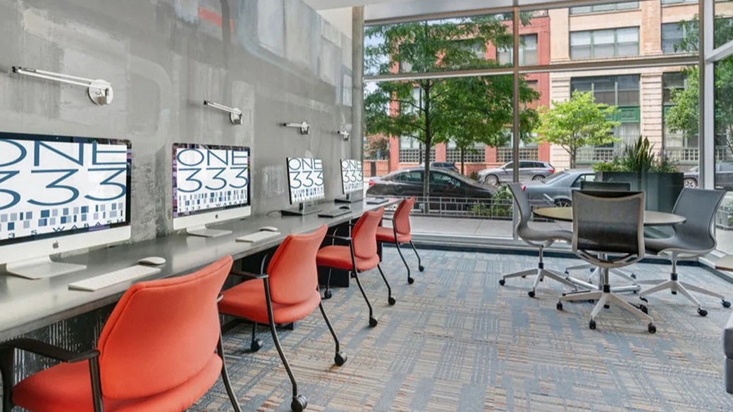 Street-level business center at ONE333 apartments in Chicago featuring iMac workstations, task lighting, and a meeting table by floor-to-ceiling windows
