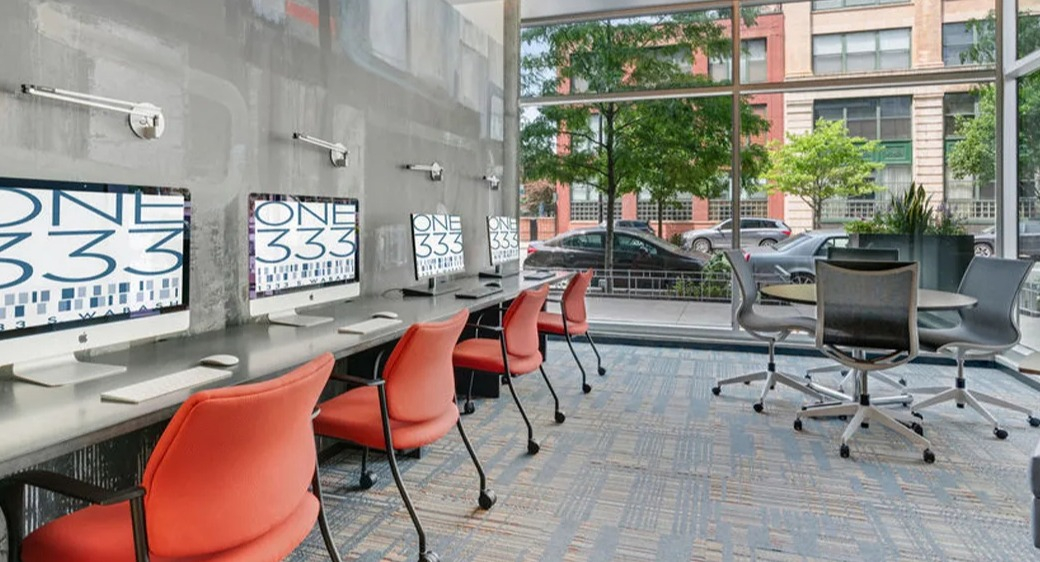 Street-level business center at ONE333 apartments in Chicago featuring iMac workstations, task lighting, and a meeting table by floor-to-ceiling windows
