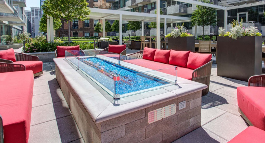 Courtyard lounge with long glass-guard fire table, bright red cushioned sofas, and pergola seating at ONE333 apartments in Chicago, ideal for relaxed evening hangouts
