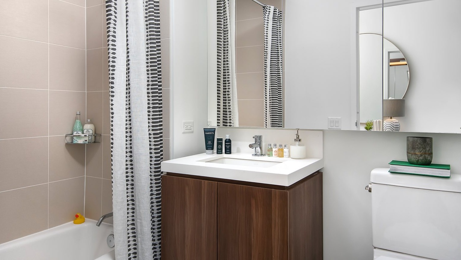 Modern apartment bathroom at OneEleven in Chicago, featuring a vanity, toilet, and a bathtub with an overhead shower and stylish curtain