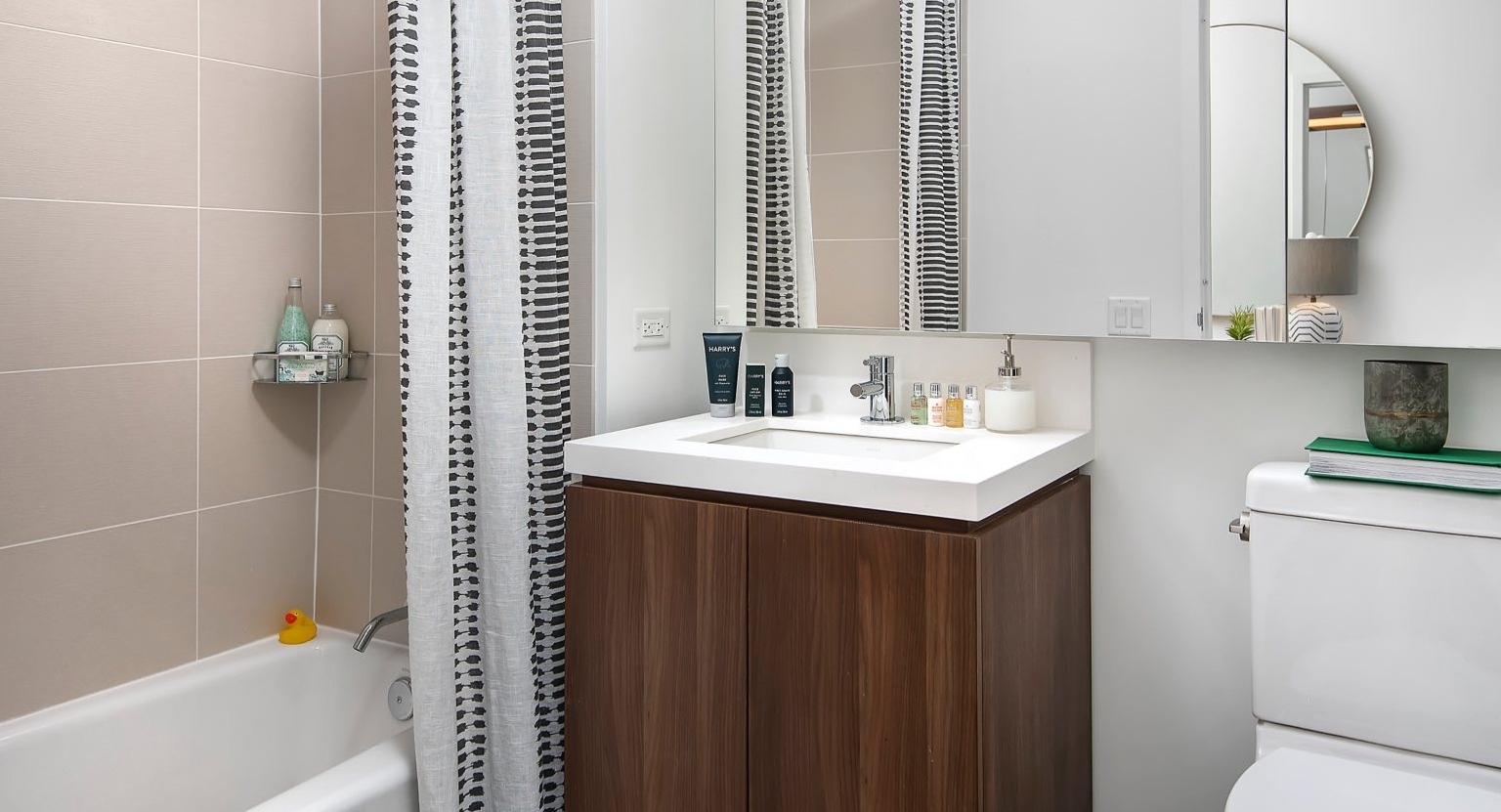 Modern apartment bathroom at OneEleven in Chicago, featuring a vanity, toilet, and a bathtub with an overhead shower and stylish curtain