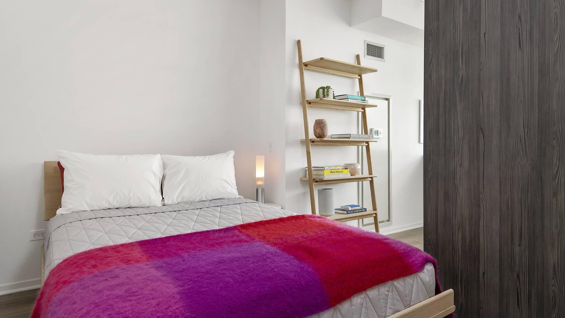 Bright bedroom at Optima Signature with minimalist decor, open shelving, and a bold red and purple blanket