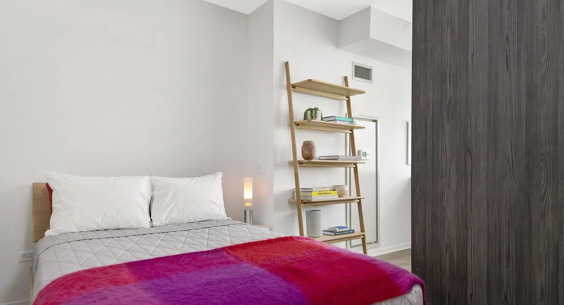 Bright bedroom at Optima Signature with minimalist decor, open shelving, and a bold red and purple blanket