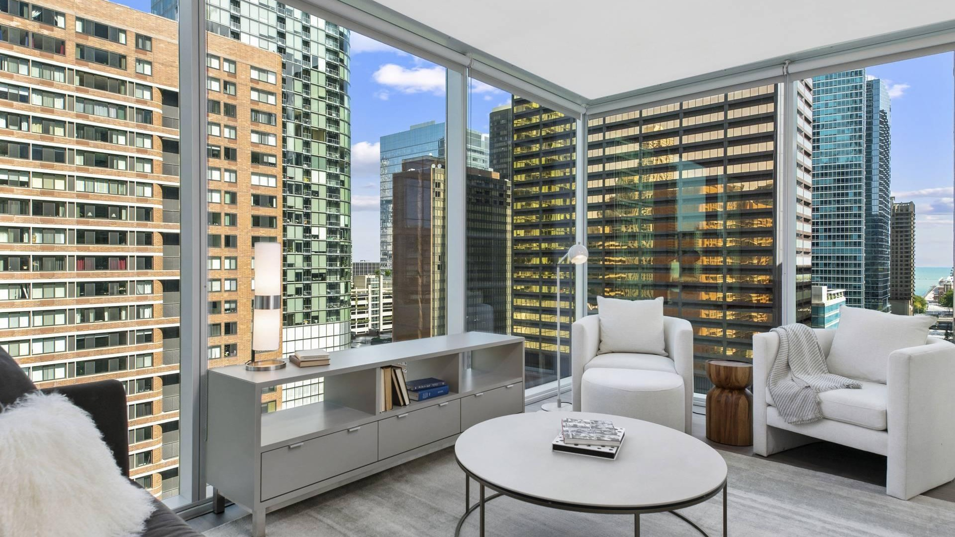 Sunlit living room at Optima Signature with sofa seating, media console, and sweeping floor-to-ceiling windows facing downtown Chicago