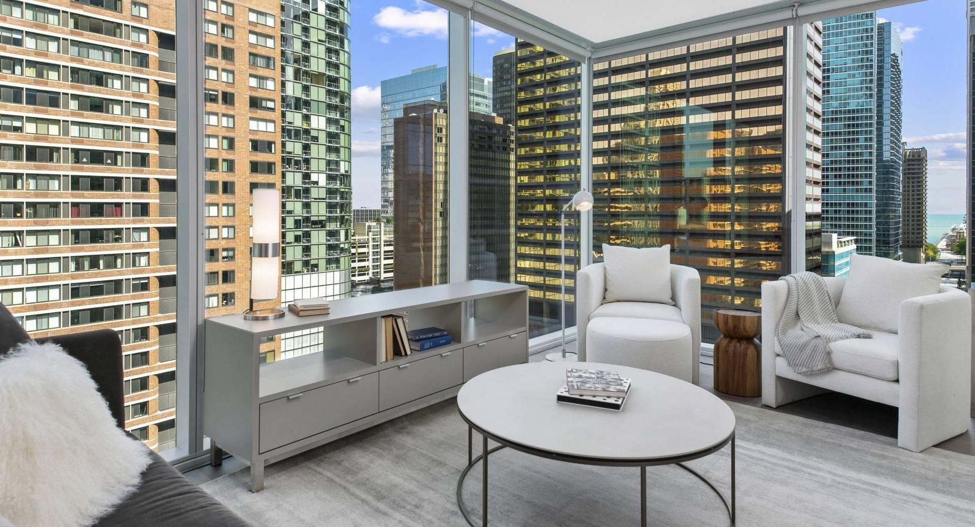 Sunlit living room at Optima Signature with sofa seating, media console, and sweeping floor-to-ceiling windows facing downtown Chicago