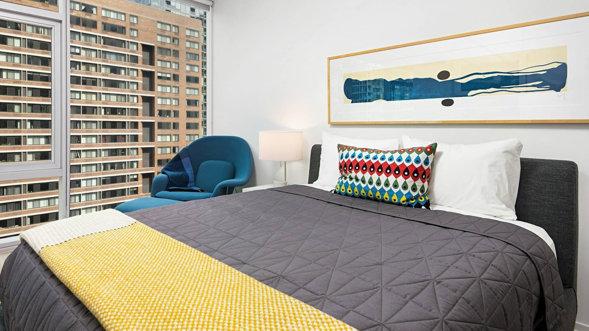 Bedroom at Optima Signature featuring a gray bed with colorful accents, blue lounge chair, and expansive windows with city views