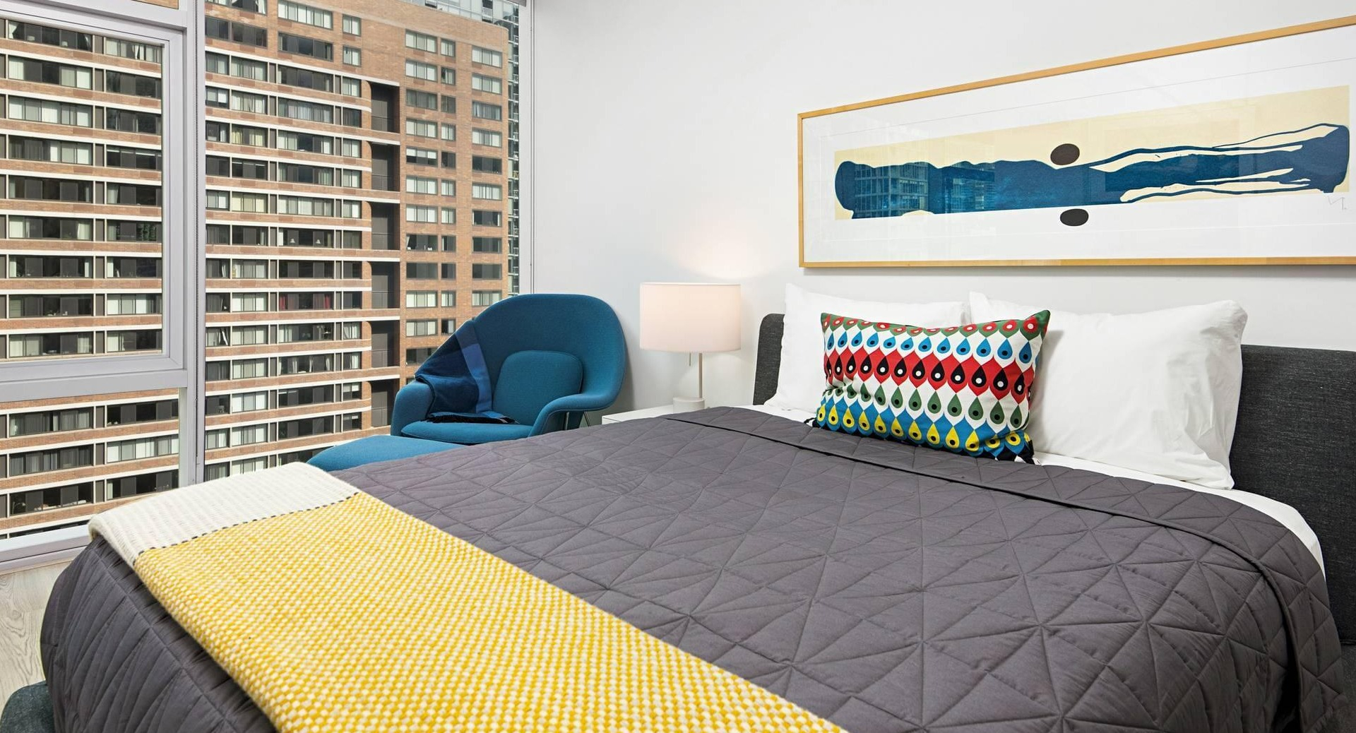 Bedroom at Optima Signature featuring a gray bed with colorful accents, blue lounge chair, and expansive windows with city views