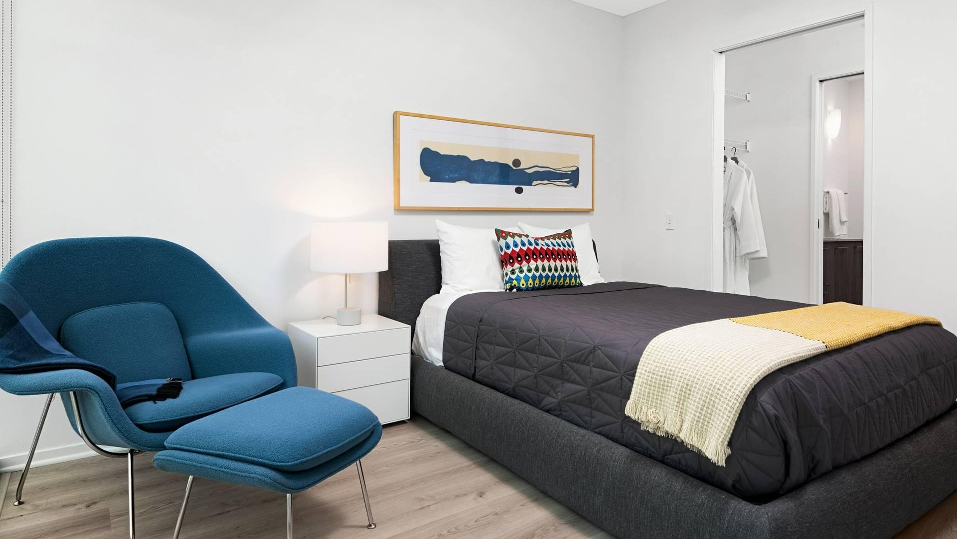 Cozy bedroom at Optima Signature with dark quilted bedding, yellow throw, and a modern lounge chair by the window