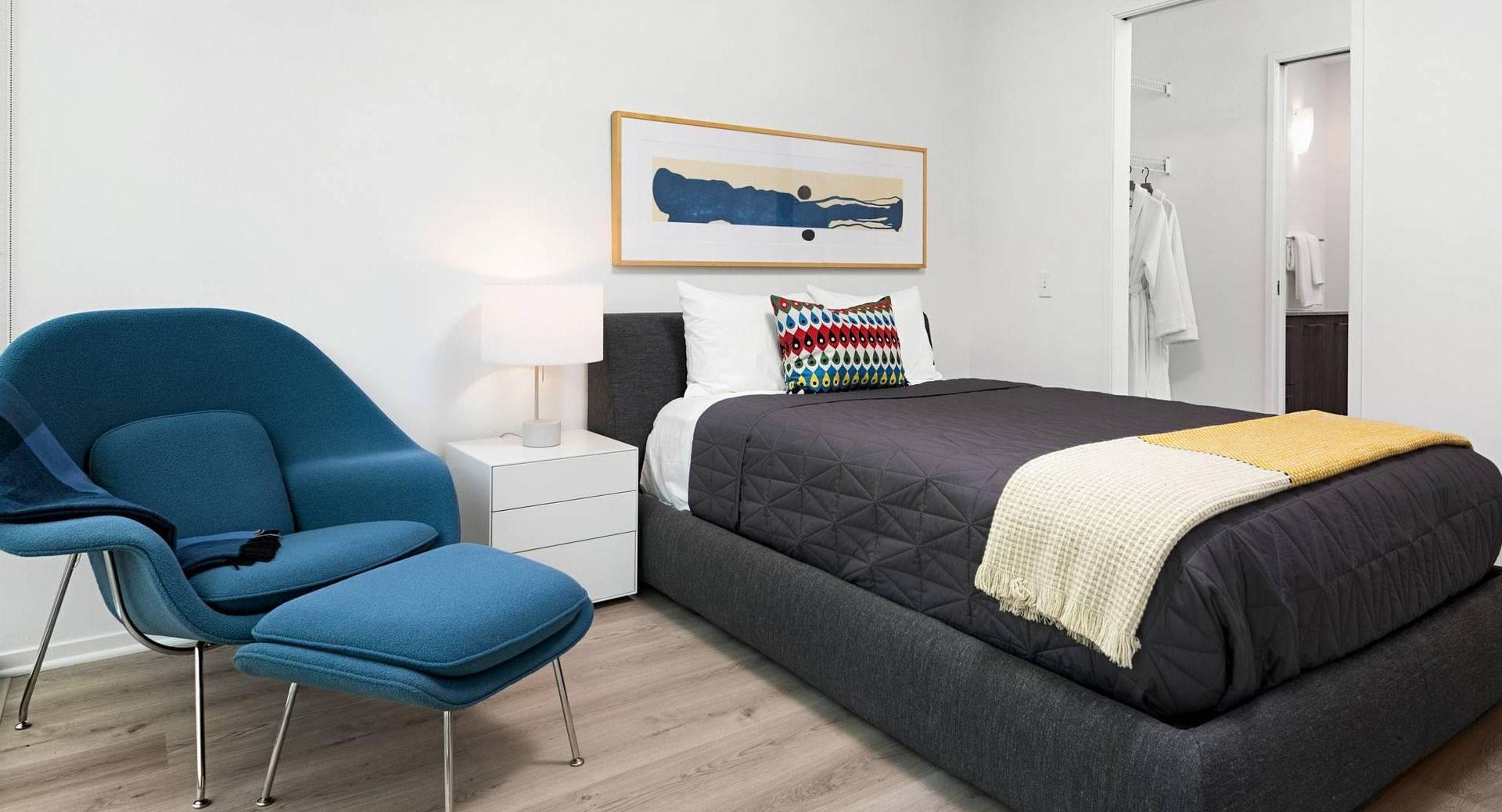 Cozy bedroom at Optima Signature with dark quilted bedding, yellow throw, and a modern lounge chair by the window