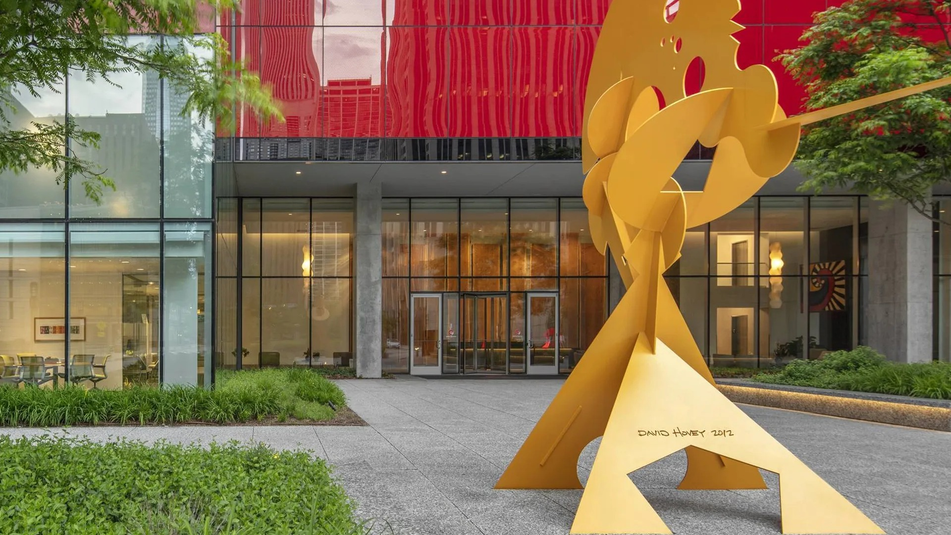Entrance plaza with red glass facade, landscaped walkway, and modern yellow sculpture at Optima Signature apartments in downtown Chicago