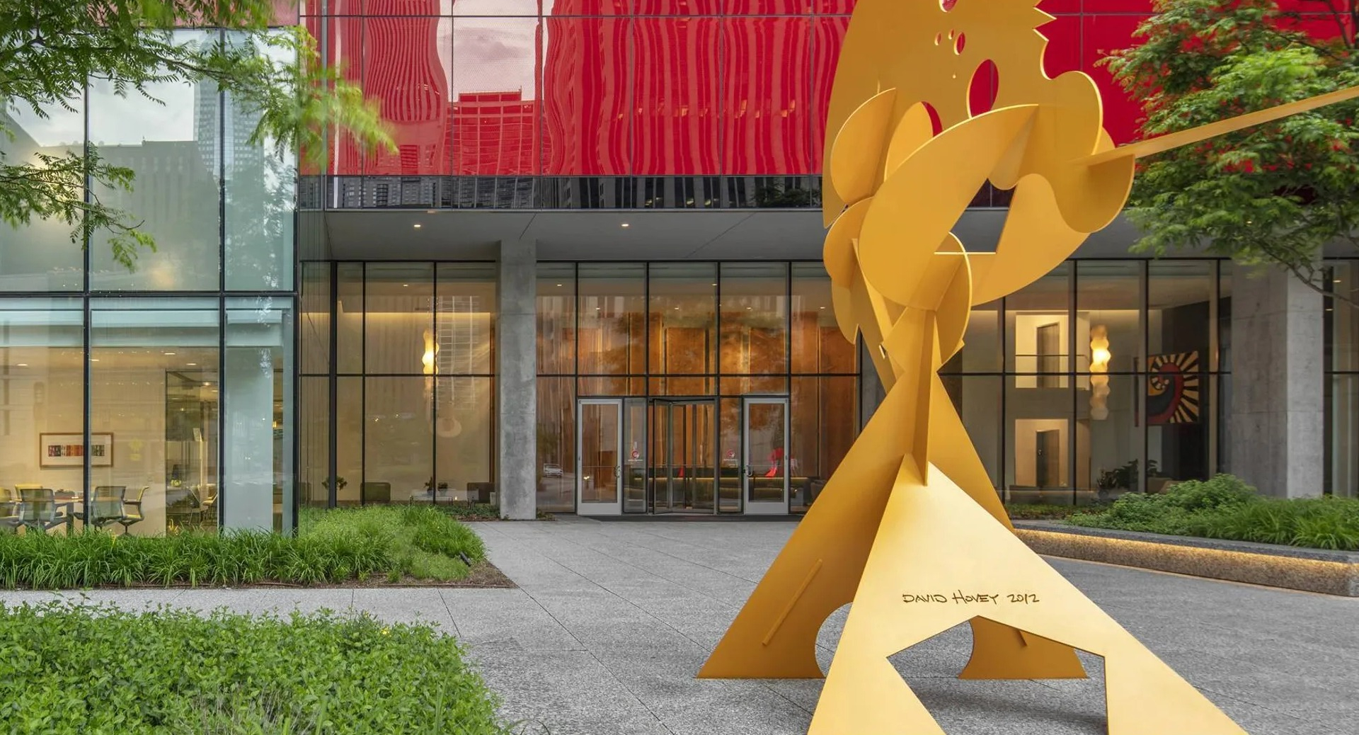 Entrance plaza with red glass facade, landscaped walkway, and modern yellow sculpture at Optima Signature apartments in downtown Chicago