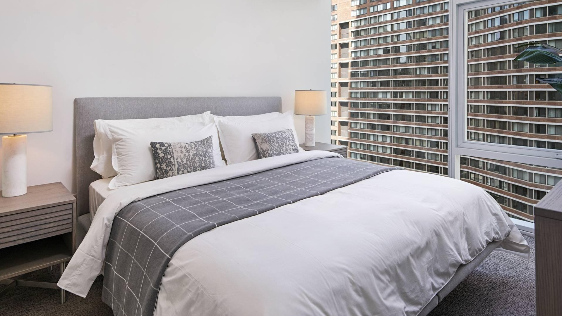 Modern bedroom at Optima Signature with floor-to-ceiling windows, gray upholstered bed, and natural light overlooking downtown Chicago