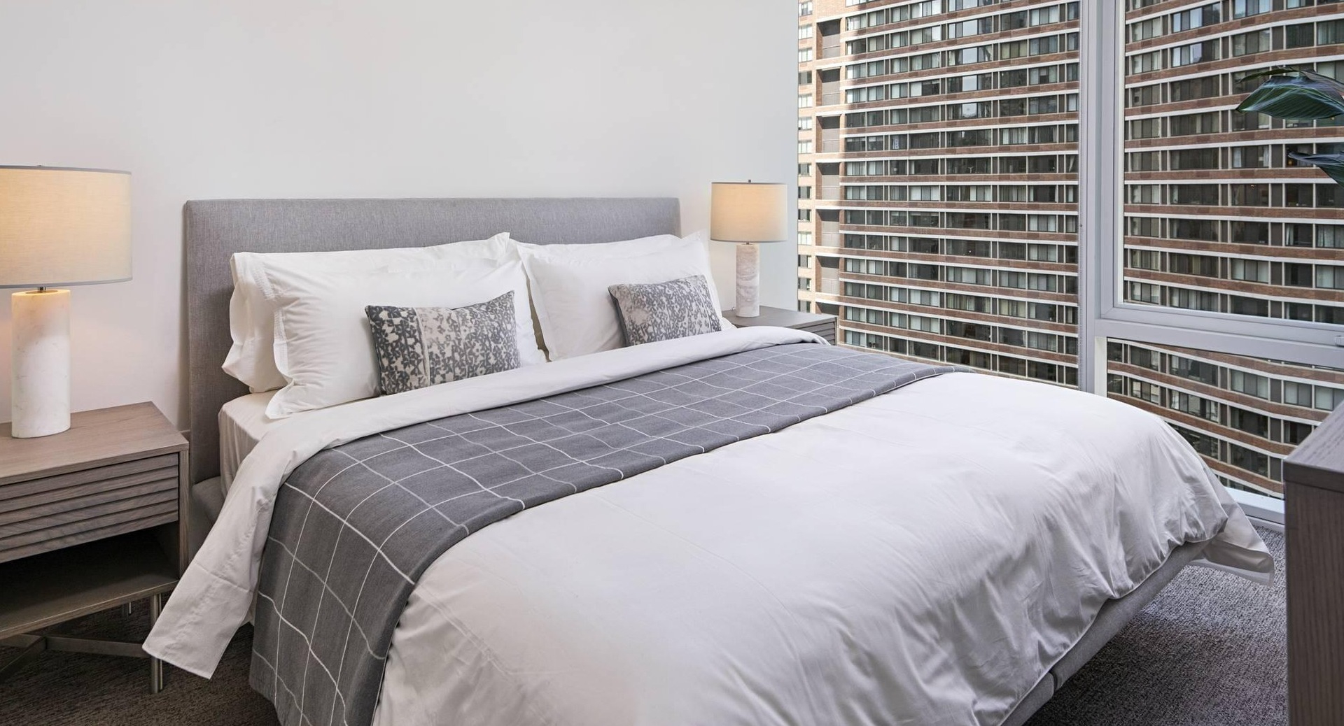 Modern bedroom at Optima Signature with floor-to-ceiling windows, gray upholstered bed, and natural light overlooking downtown Chicago