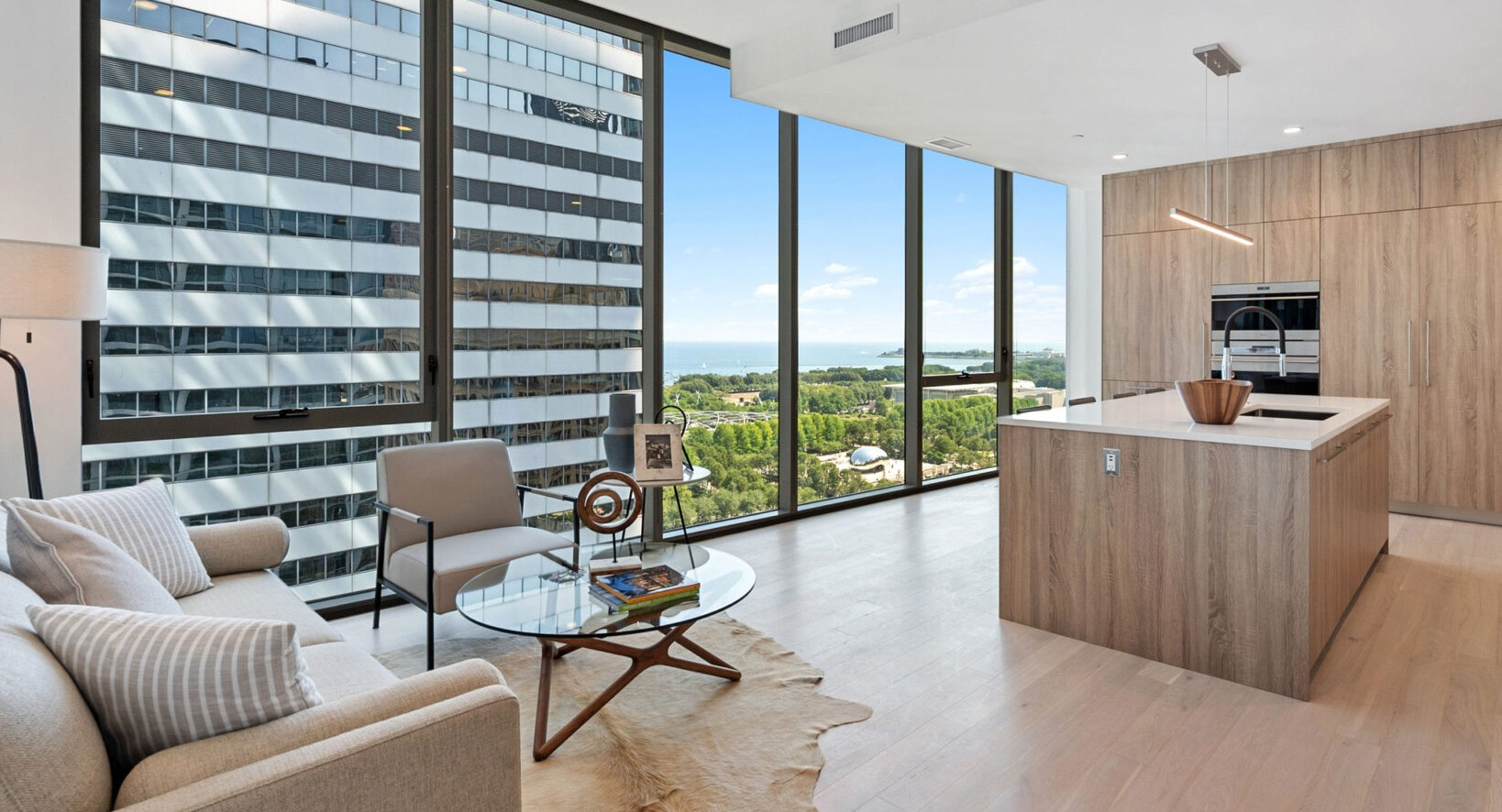 Bright open-plan living room and kitchen with wood cabinetry, a large island, and expansive windows offering city and lake views at Parkline Chicago in Chicago