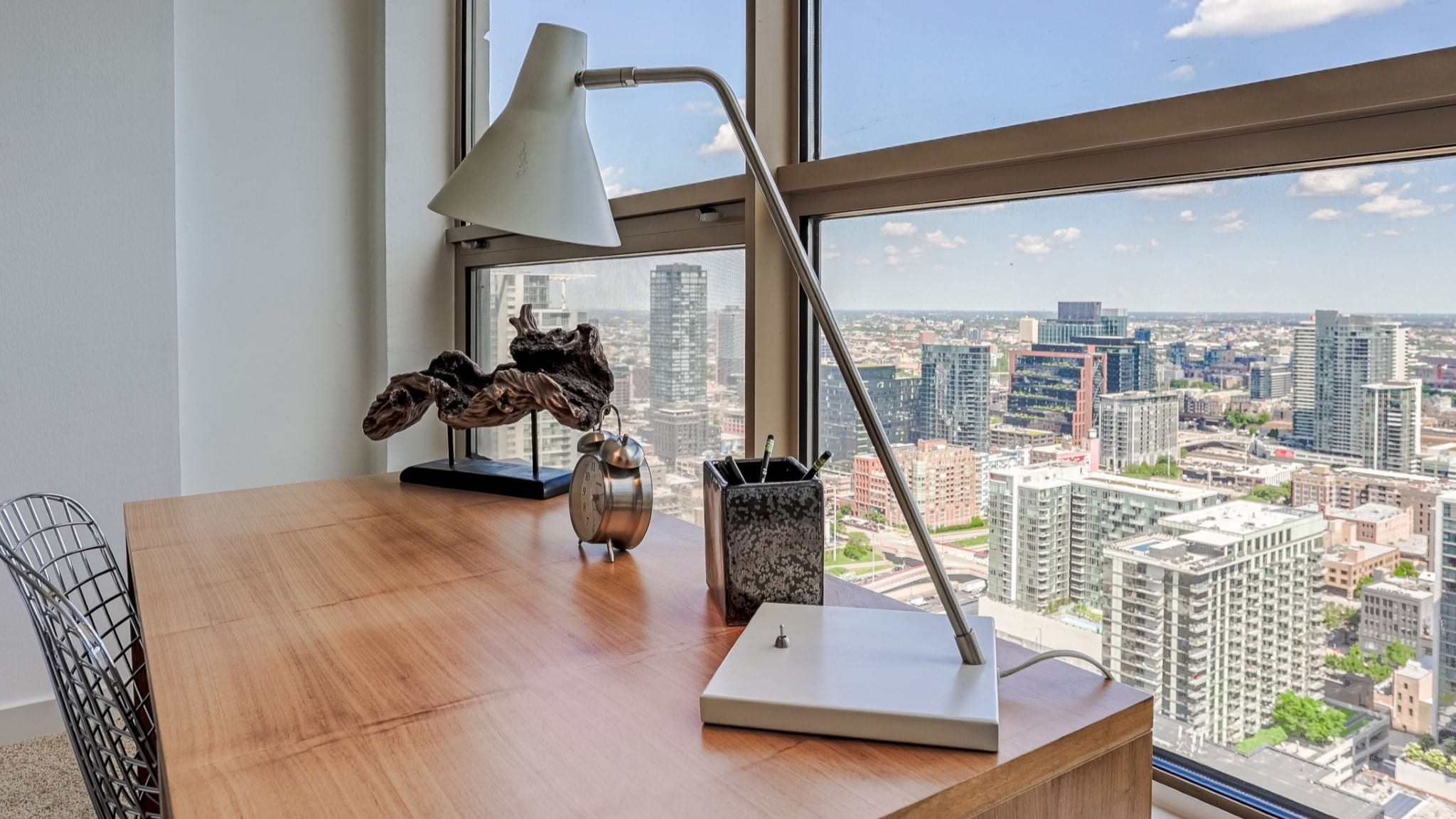 Bright bedroom at Presidential Towers in Chicago showcasing a wood desk by expansive windows overlooking the city, plus modern decor and a sleek lamp