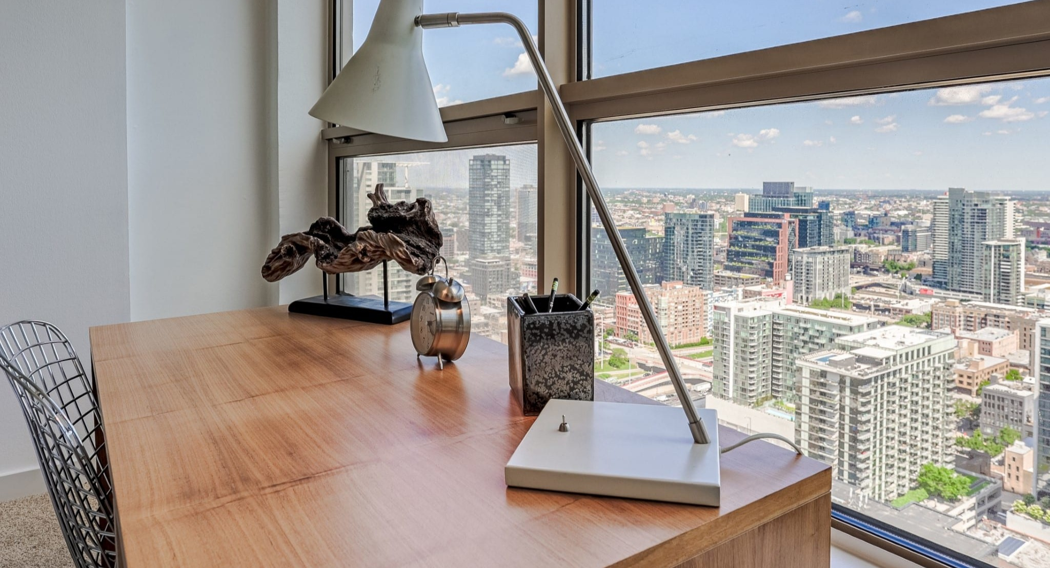 Bright bedroom at Presidential Towers in Chicago showcasing a wood desk by expansive windows overlooking the city, plus modern decor and a sleek lamp