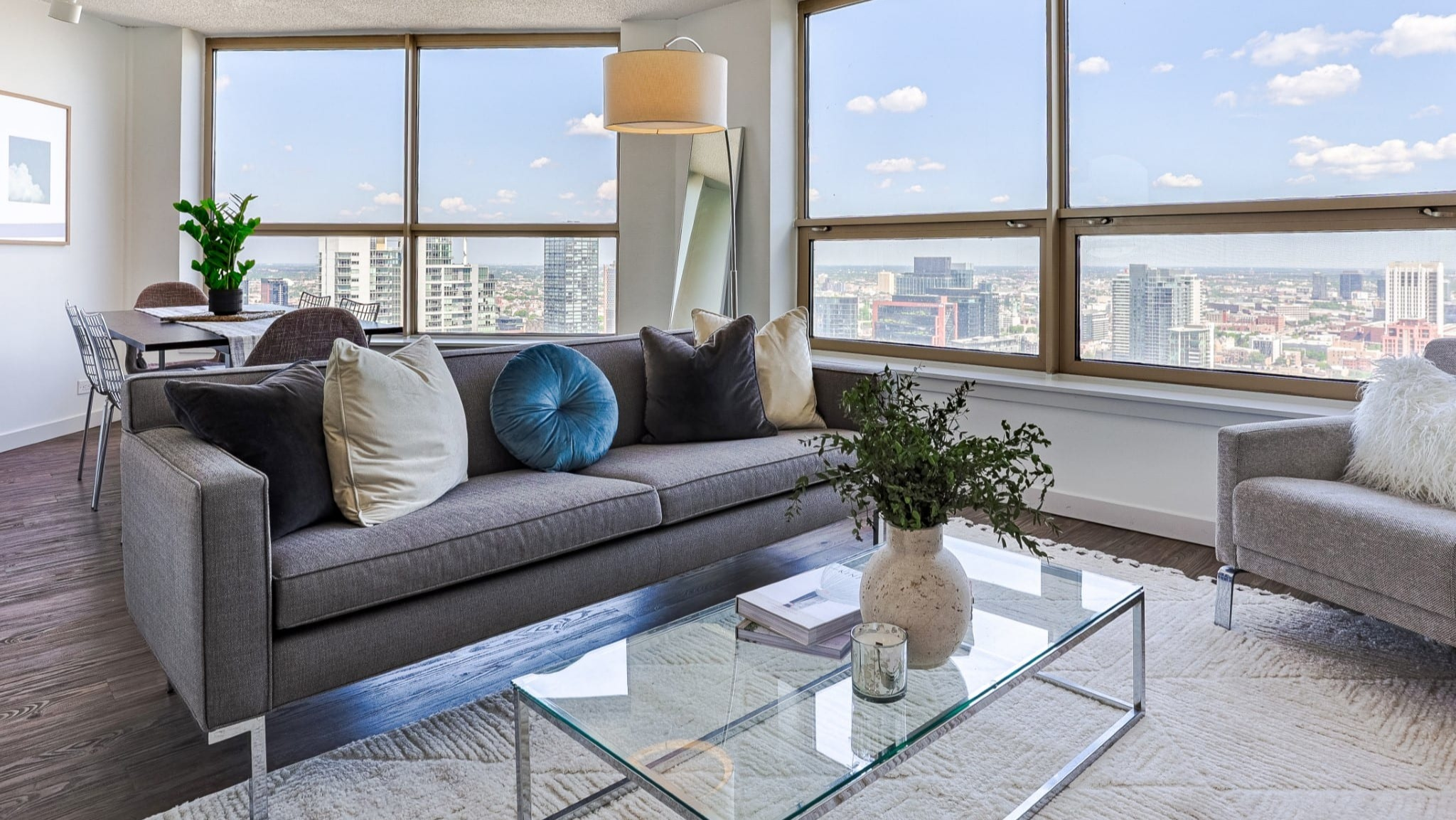 Sunlit corner living room at Presidential Towers in Chicago with wide windows, gray sofa, armchair, glass coffee table, and expansive skyline views