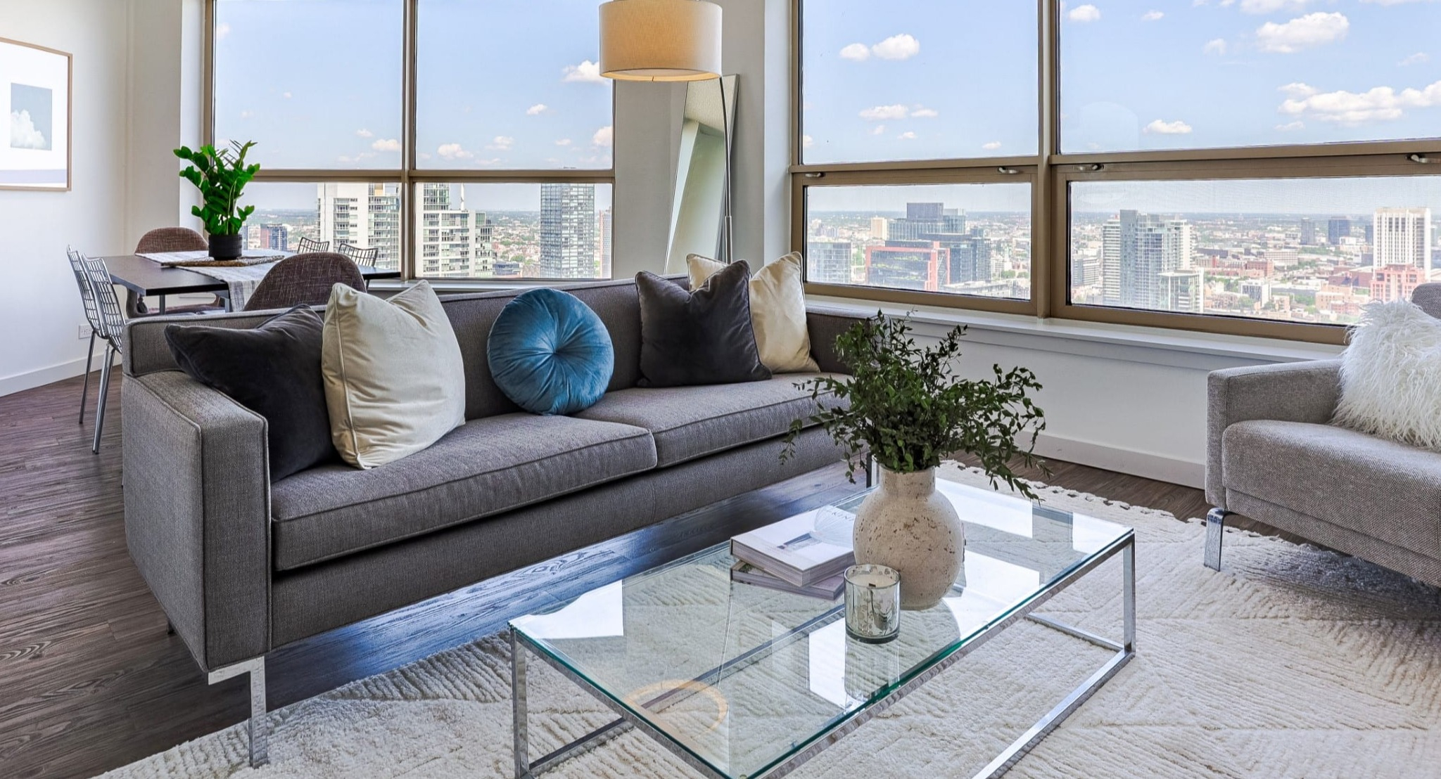 Sunlit corner living room at Presidential Towers in Chicago with wide windows, gray sofa, armchair, glass coffee table, and expansive skyline views