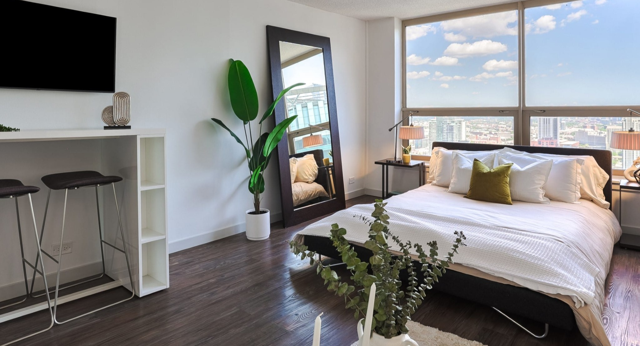 Cozy studio sleeping area at Presidential Towers in Chicago with low platform bed, large windows, city views, and a tall mirror beside a leafy plant