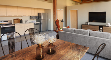A stylish living room and kitchen area featuring exposed wooden beams, modern furniture, and a rustic dining table at River West Lofts in Chicago