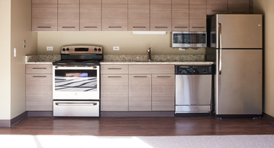 A modern kitchenette featuring light wood cabinets, granite countertops, stainless steel appliances, and ample space at River West Lofts in Chicago