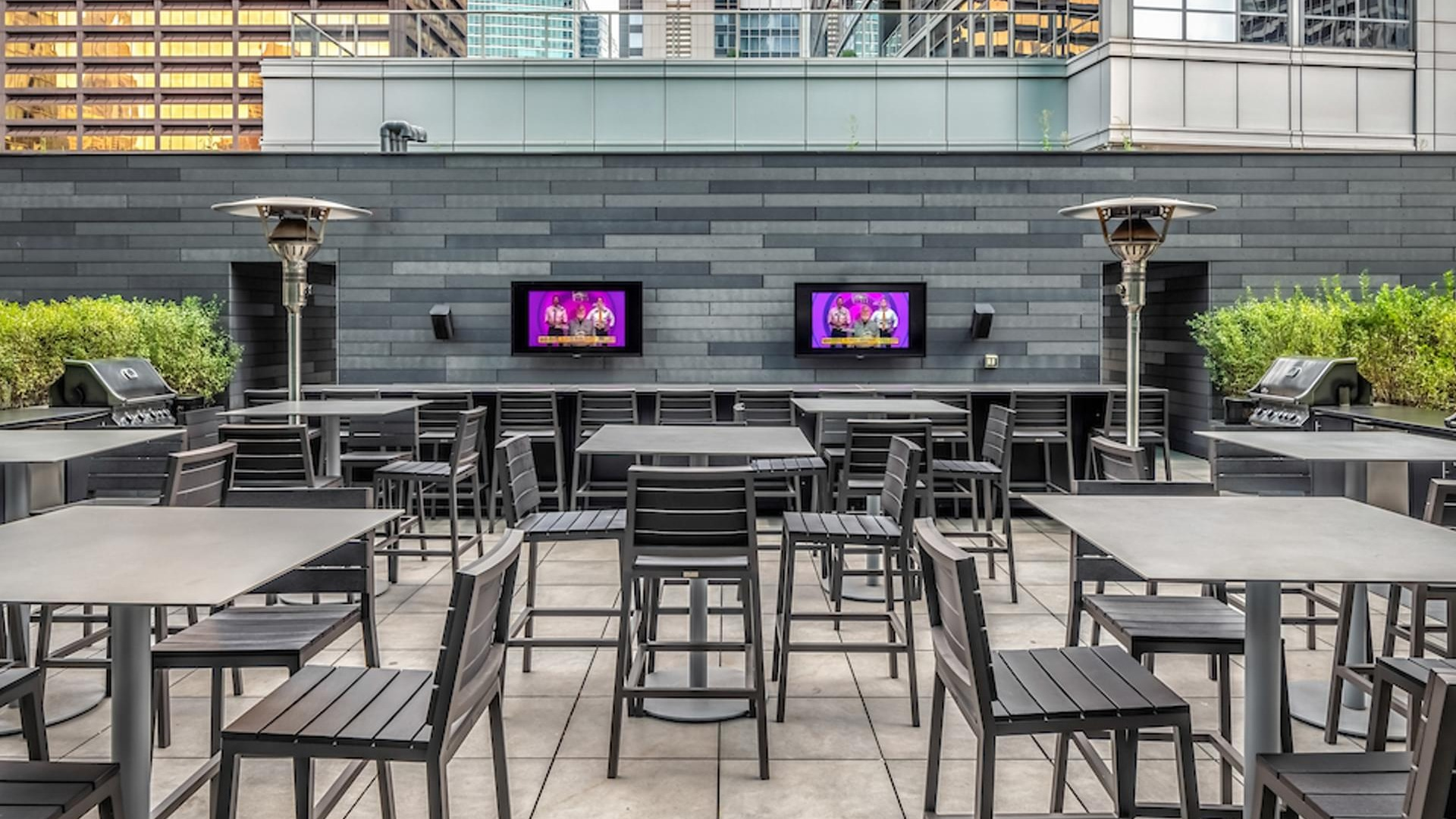 Rooftop grilling lounge with dining tables, TVs, and stainless-steel BBQ stations surrounded by city views at Optima Signature Chicago
