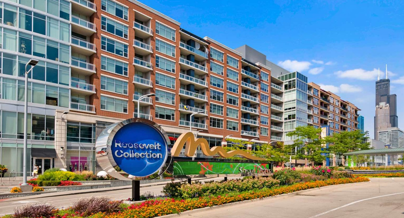 Exterior view of Roosevelt Collection Lofts with a prominent sign, lush landscaping, and a bustling urban setting