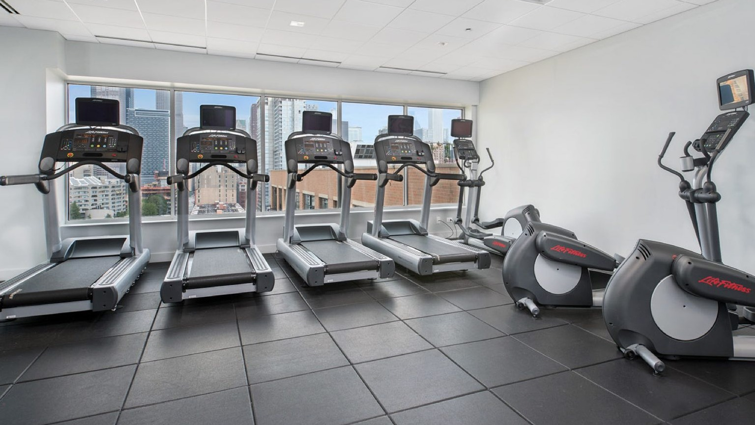 Modern gym with a selection of treadmills, ellipticals, and other cardio equipment, offering city views from windows at Roosevelt Collection Lofts