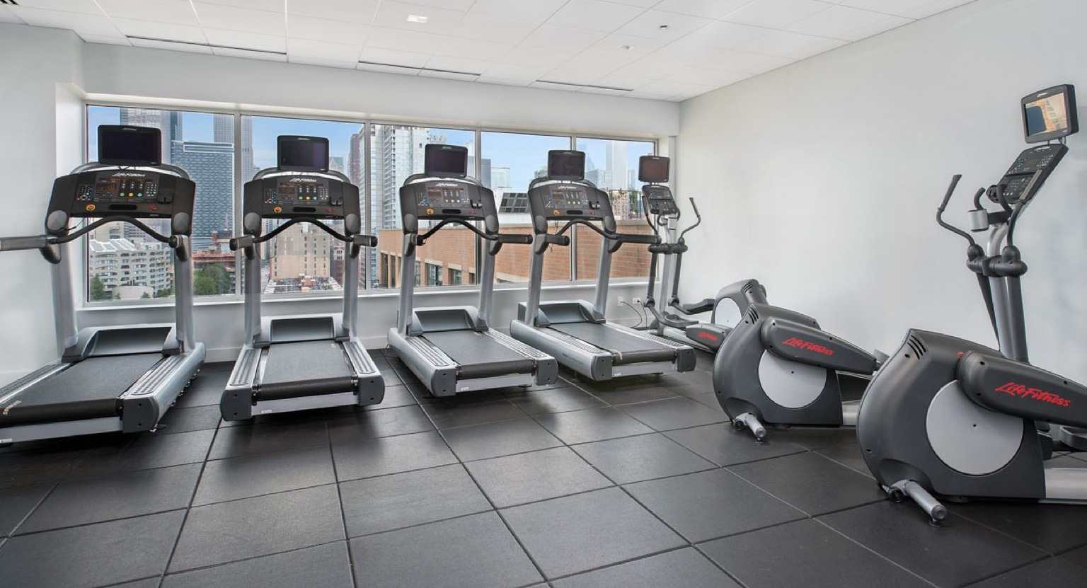 Modern gym with a selection of treadmills, ellipticals, and other cardio equipment, offering city views from windows  at Roosevelt Collection Lofts