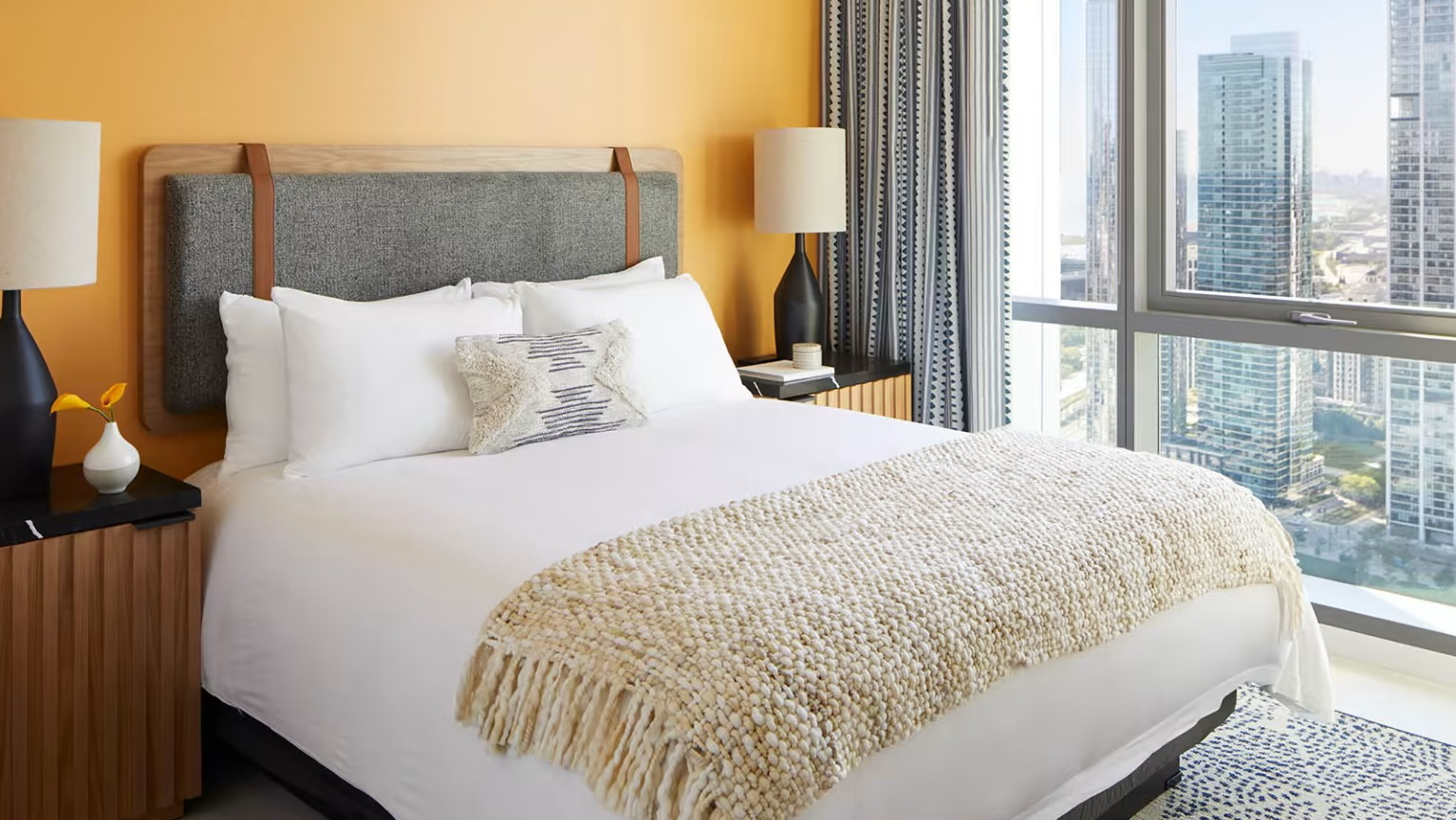 Bright and cozy bedroom at Sentral Michigan Avenue in Chicago, featuring a comfortable bed, warm orange wall, and stunning city skyline views