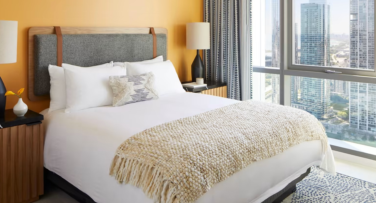 Bright and cozy bedroom at Sentral Michigan Avenue in Chicago, featuring a comfortable bed, warm orange wall, and stunning city skyline views