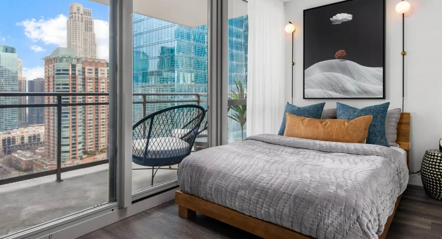 Cozy bedroom at Shoreham and Tides apartments in Chicago with floor-to-ceiling glass, private balcony seating, and modern bed styling