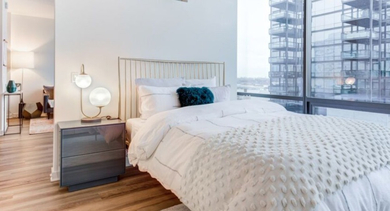 A bright bedroom at SoNo East Apartments in Chicago, featuring a comfortable bed and expansive city views from large windows