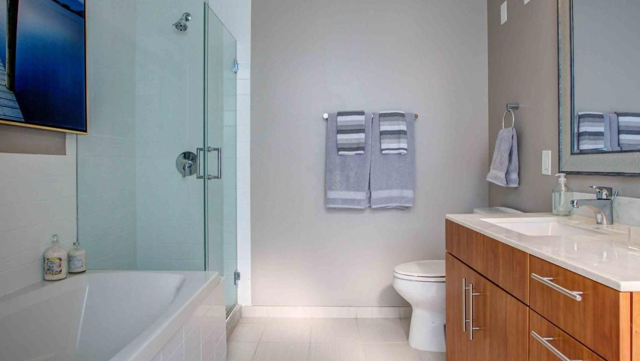 Modern bathroom featuring a deep soaking tub with a glass shower enclosure, wood vanity, and ample lighting at State and Grand Apartments in Chicago