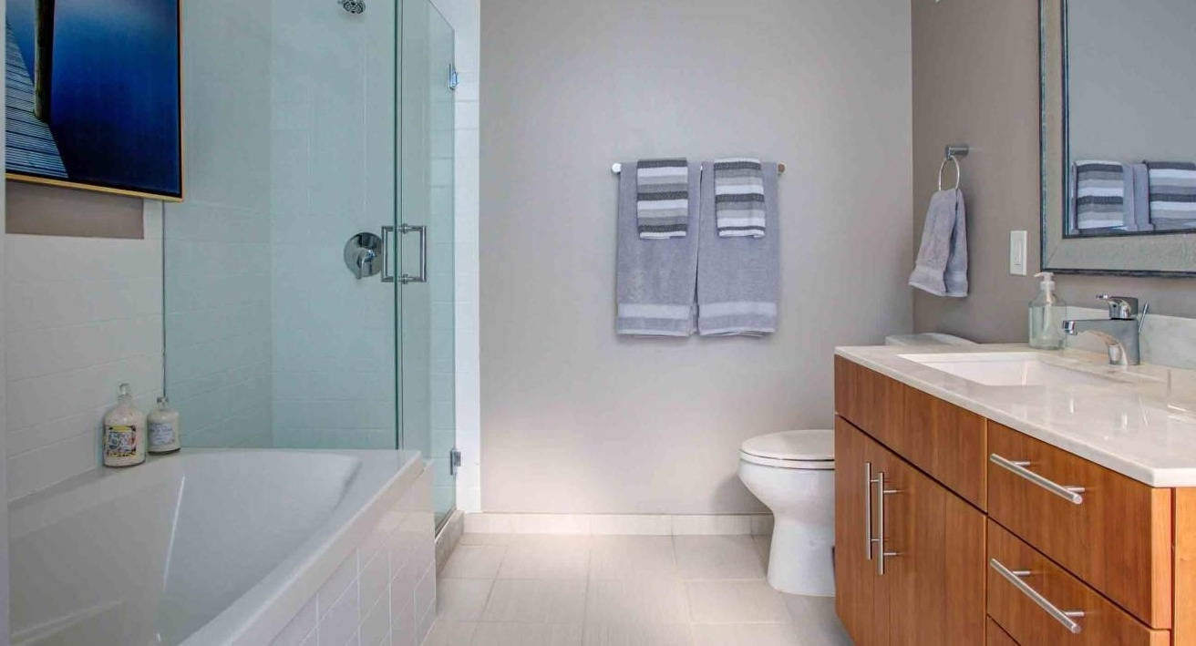 Modern bathroom featuring a deep soaking tub with a glass shower enclosure, wood vanity, and ample lighting at State and Grand Apartments in Chicago