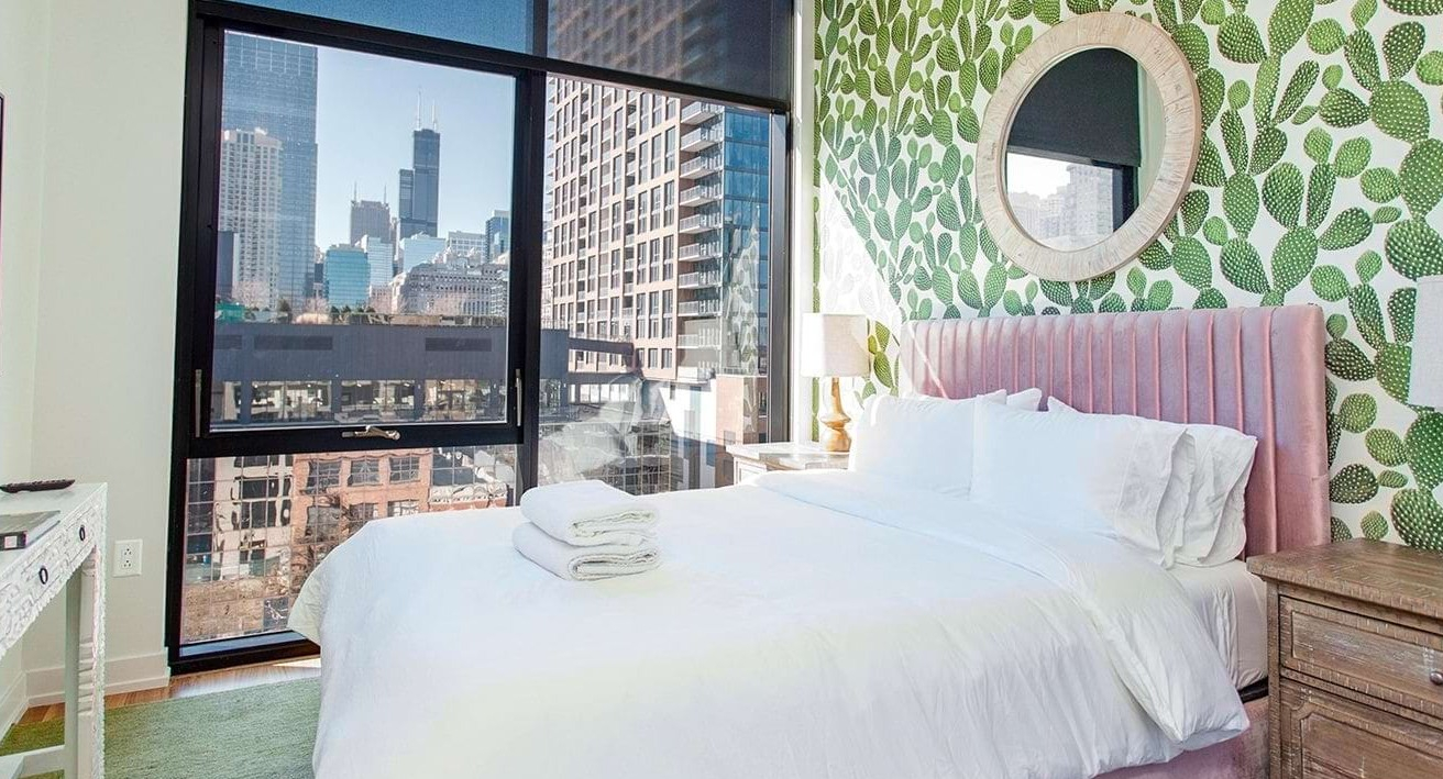 Cozy bedroom at The Ardus in Chicago, featuring a comfortable bed, vibrant cactus wallpaper, and expansive city views through large windows